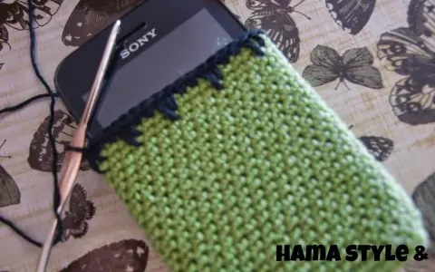 Funda de móvil tejida a crochet, un patrón verde con borde negro. Un gancho y hilo negro están listos para continuar el proyecto.