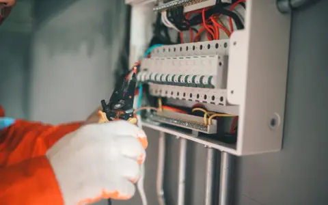 Elektryk w białych rękawicach wywala bezpiecznik w skrzynce, używając szczypiec.