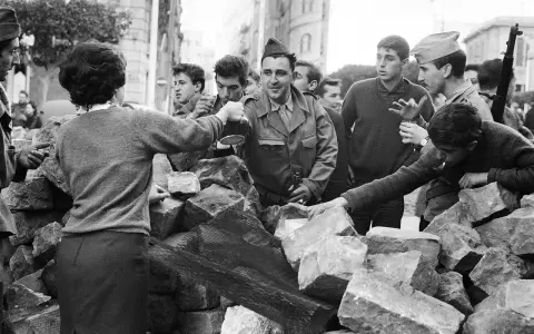 Un grupo de jóvenes y soldados interactúan tras una barricada de piedras en Argelia. Un hombre con uniforme militar sirve bebida.