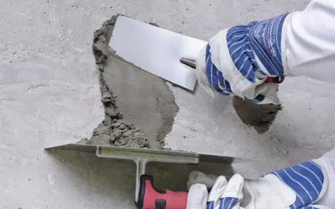 Naprawa betonu na zewnątrz. Ręce w rękawicach nakładają zaprawę kielnią na uszkodzony fragment ściany.
