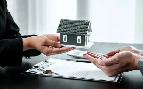 Manos entregan maqueta de casa sobre contrato y llaves. Posible exención IRPF venta vivienda heredada.