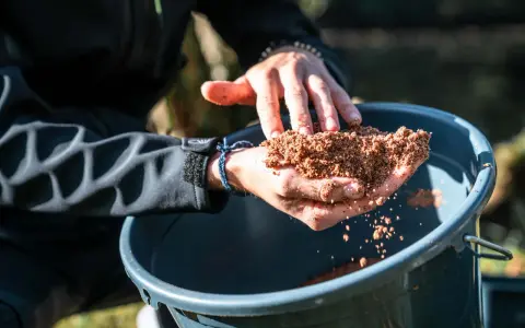 Ręce trzymają zanętę Traper Feeder nad wiaderkiem.