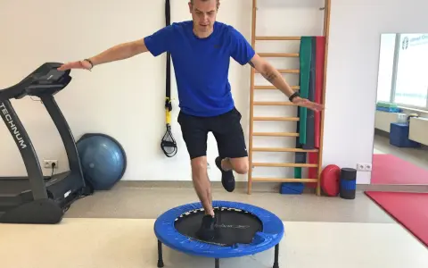 Mężczyzna wykonuje ćwiczenia po rekonstrukcji ACL, balansując na małej trampolinie. W tle bieżnia i drabinka gimnastyczna.