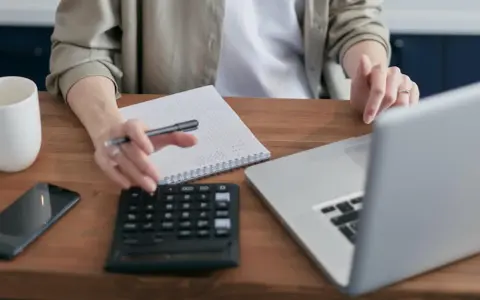 Persona revisa cuentas en su portátil, calculadora y libreta. Podría estar gestionando la hacienda pública deudora por IVA.