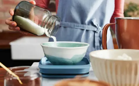Eine Hand schüttet Mehl aus einem Glas in eine Schüssel. Die Frage, wie viel Gramm Mehl in eine Tasse passen, wird hier visuell thematisiert.