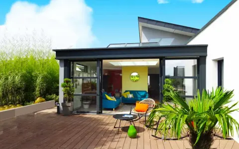 Une véranda moderne avec de grandes baies vitrées s'ouvrant sur une terrasse en bois. Un salon coloré est visible à l'intérieur.