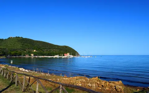 A tranquil bay in Tuscany with sailboats docked, a green hillside, and a rustic wooden fence in the foreground.