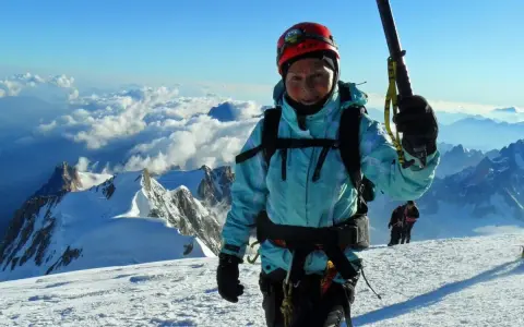 Barbara Prymakowska, mimo swojego wieku, zdobywa szczyty. Uśmiechnięta alpinistka w kasku i turkusowej kurtce na tle gór i chmur.