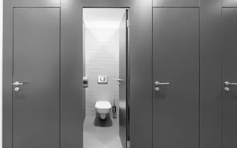 Reihe von grauen Türen zu Toilettenkabinen. Eine Tür ist geöffnet und gibt den Blick auf eine Toilette frei. Gute Frage, wie viele Toiletten pro Mitarbeiter?