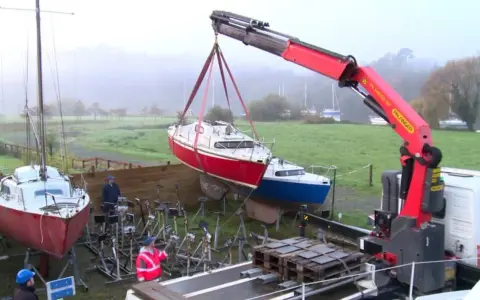 Grue soulevant un bateau rouge pour son recyclage. Un autre bateau bleu est posé sur des supports. L'opération de recyclage bateau se déroule dans un paysage brumeux.