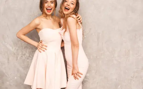 Dwie uśmiechnięte kobiety w pastelowych sukienkach, idealne na różne rodzaje dress code'u, od koktajlowego po wieczorowy.