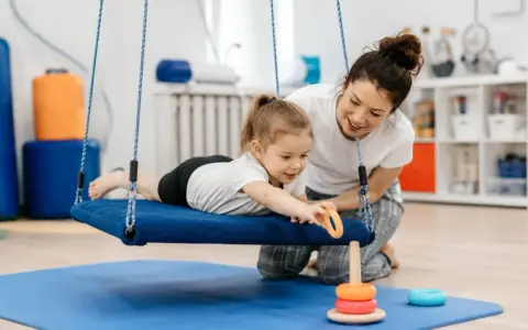 Terapia integracji sensorycznej: dziewczynka na huśtawce z terapeutką, która pomaga jej sięgnąć po zabawkę.