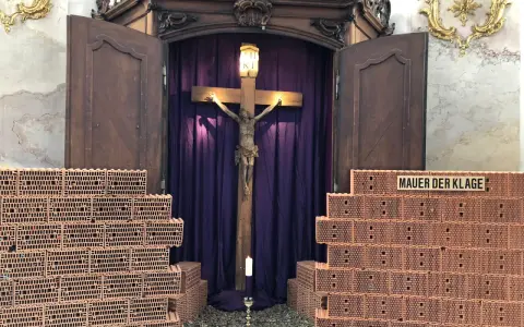 Kreuzweg Stationen: Jesus am Kreuz, umgeben von Ziegelmauern, die die "Mauer der Klage" symbolisieren. Eine Kerze brennt vor dem Kruzifix.