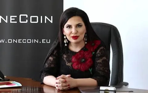 Rupert Ignatov, w czarnej koronkowej sukni z czerwonymi kwiatami, siedzi przy biurku.