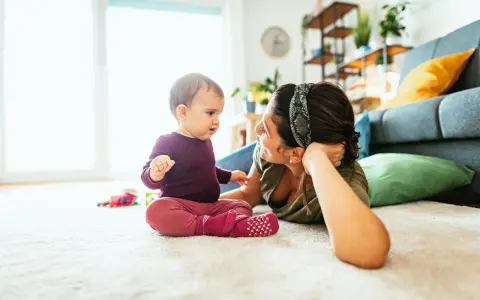 Mama patrzy na dziecko, które siedzi na dywanie. To obrazek pokazujący, co powinien umieć 2 latek: rozwijać się i bawić.