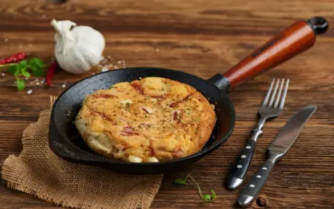 Pyszny omlet wytrawny z kawałkami wędliny, podany na patelni. Obok widelec i nóż.