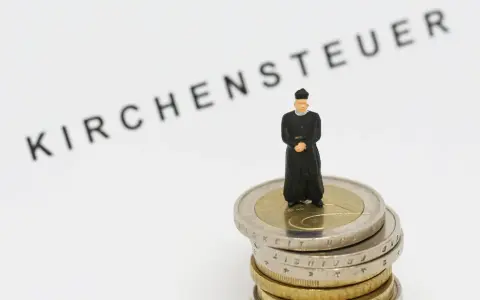 Miniatur-Priester steht auf Münzstapel. Der Schriftzug "Kirchensteuer" im Hintergrund wirft die Frage auf, warum man kirchensteuer zahlt.
