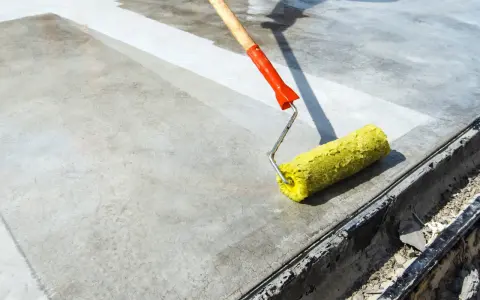Wałek z żółtym włosiem rozprowadza środek do impregnacji betonu na szarej powierzchni.