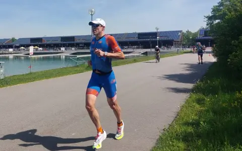 Triathlet im blauen Anzug läuft auf einer Straße. Er fragt sich, wie wird man schneller. Ein Radfahrer ist im Hintergrund zu sehen.