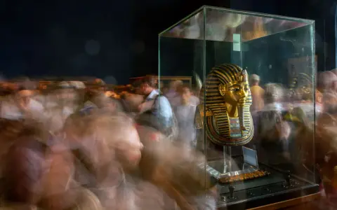 Goldene Maske des Pharao Tutanchamun in einer Vitrine, umgeben von verschwommenen Besuchern.
