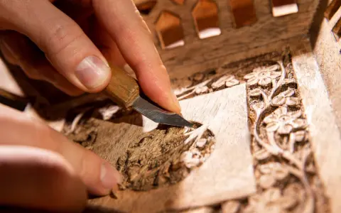 Main d'artisan sculptant un motif floral sur un meuble en bois, étape clé pour savoir comment nettoyer un meuble en marqueterie.