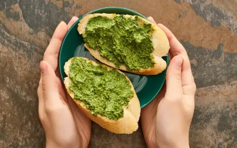 Dwie kromki bagietki z aromatycznym pesto z pietruszki na zielonym talerzyku, trzymanym w dłoniach.