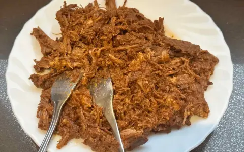 Delicioso pulled pork deshebrado en un plato blanco, listo para ser servido. Dos tenedores se clavan en la carne jugosa.