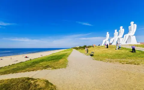 De vita sandstränderna i Danmark möter en grönskande sluttning med gigantiska sittande figurer. En grusväg leder mot havet under en klarblå himmel.