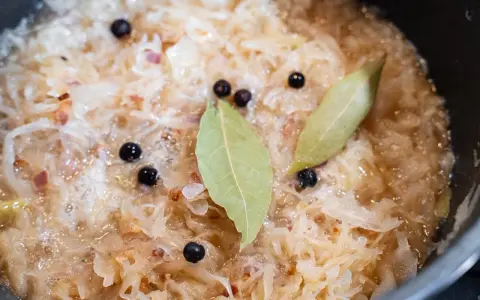 Sauerkraut köchelt mit Lorbeerblättern und Wacholderbeeren, um den Geschmack zu verfeinern. Kleine rote Zwiebelstücke sind ebenfalls sichtbar.
