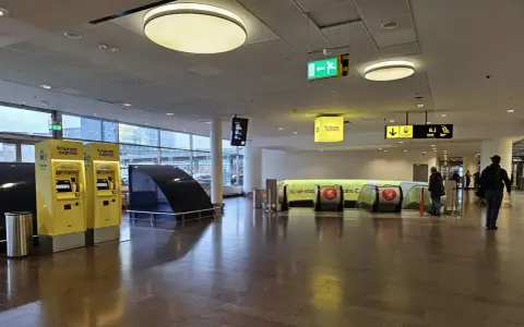 Żółte automaty biletowe Arlanda Express na lotnisku w Sztokholmie. W tle ruchome schody i znaki kierujące.