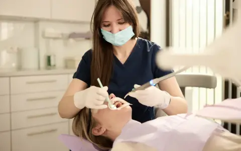 Dentystka w masce i rękawiczkach wykonuje zabieg u pacjentki w gabinecie stomatologicznym.