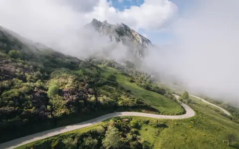 Strada tortuosa che sale verso le vette nebbiose, evocando l'ascesa al **Col du Tourmalet**.