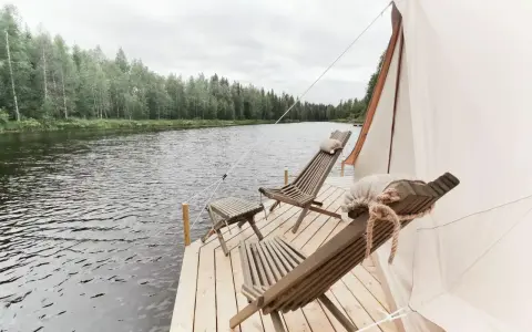 Två träfåtöljer och ett litet bord på en brygga vid en sjö, med skog i bakgrunden. En del av ett glampingtält syns. Upplev glamping Norrbotten.