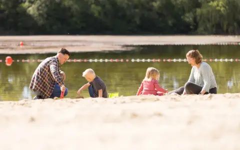 Familie spielt am Sandstrand des badesee mindenerwald. Vater hilft Sohn beim Sandburgen bauen, während Mutter und Tochter daneben sitzen.