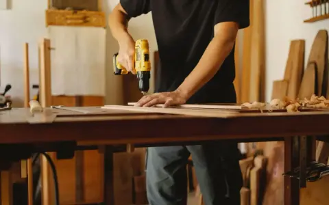 Mann bohrt Holzbretter, um seine Werkstatt einzurichten. Hobelspäne liegen auf dem Tisch.
