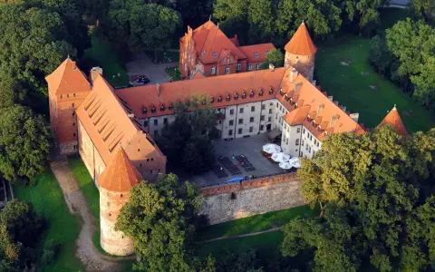 Zamek w Bytowie otoczony zielenią, z czerwonymi dachami i okrągłymi wieżami. Na dziedzińcu widać stoliki i parasole.