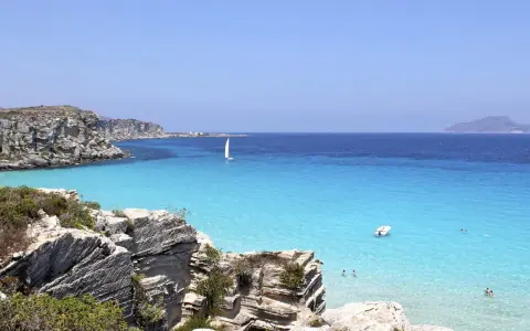 Scogliere rocciose e mare cristallino a Favignana. Un veliero solca le acque turchesi, perfetto per scoprire cosa vedere a Favignana in un giorno.