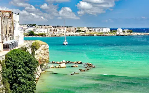 Vista mozzafiato del paese di Otranto con le sue case bianche affacciate sul mare turchese e un veliero solitario.
