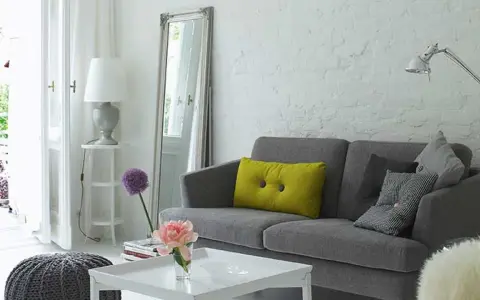 Sofá gris con cojines, espejo alto y lámpara. Trucos para decorar espacios pequeños con estilo y amplitud.