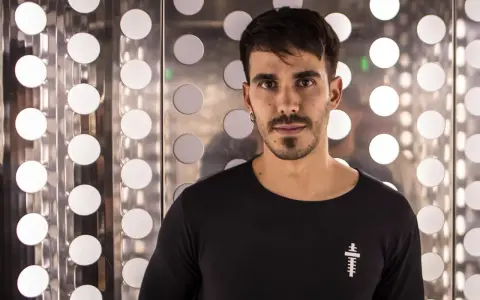 Raúl Pacheco, DJ, posa con una camiseta negra frente a un fondo metálico con círculos blancos brillantes.
