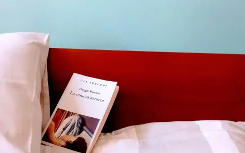 Un libro aperto, "La camera azzurra" di Simenon, giace su un letto bianco, pronto per essere letto.