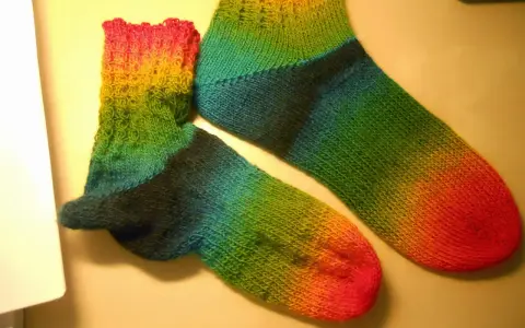 Två regnbågsfärgade, handstickade sockor i ett stycke. En större och en mindre, med färger som går från rosa och gult till grönt och blått.