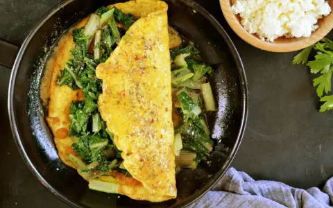 Omlet z serka wiejskiego z zielonymi warzywami na patelni. Obok miseczka z serkiem i natka pietruszki.
