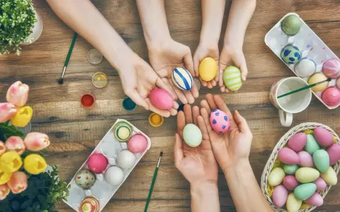 Hände von Kindern und Erwachsenen bemalen bunte Ostereier. Ein Korb voller Eier und Tulpen auf Holztisch.