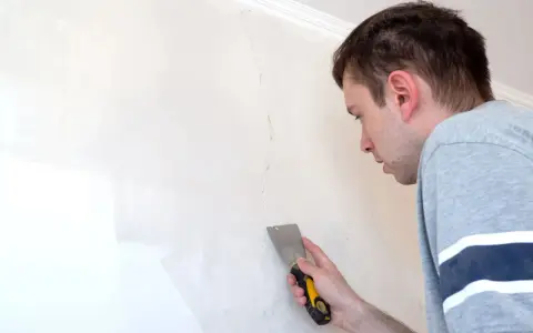 Uomo con spatola ripara una crepa su un muro bianco.