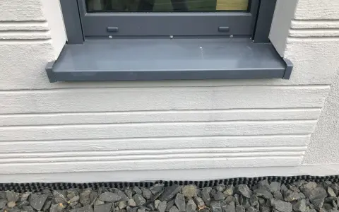 Graue Fensterbank mit leichter Neigung, die Regenwasser ableitet. Darunter weiße Holzverkleidung und grauer Schotter.