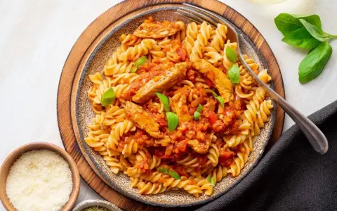 Pyszny makaron z kurczakiem w sosie pomidorowym, udekorowany bazylią. Obok miseczki z parmezanem i ziołami.