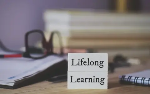 Dwa białe klocki z napisami "Lifelong" i "Learning" symbolizują uczenie się przez całe życie. W tle okulary i notatnik.