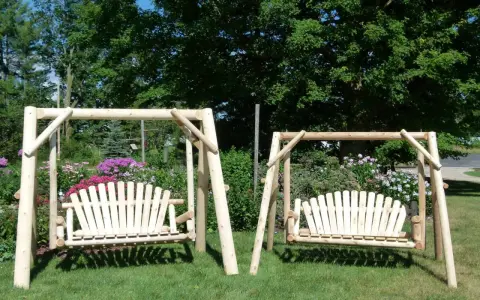Two wooden porch swings stand on a grassy lawn, surrounded by colorful flowers and lush trees. This image offers inspiration for how to hang a porch swing.