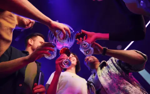 Grupa przyjaciół wznosi toast, świętując weekend. W tle dyskotekowe światła, sugerujące zabawę i być może początek weekendowego alkoholizmu.